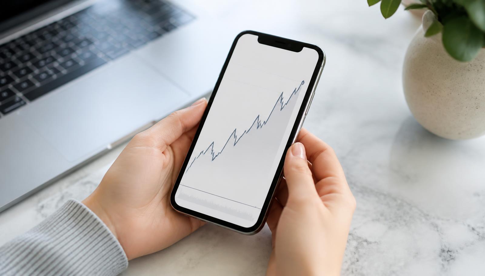 Hand hält Smartphone mit neutralem Portfolio-Chart über einem hellen Schreibtisch