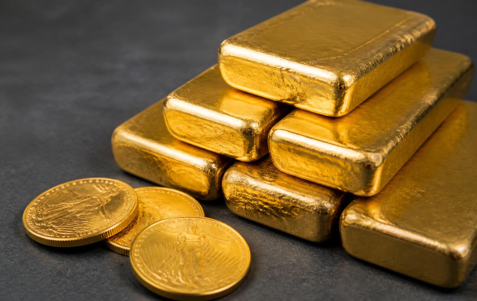 Gestapelte Feingoldbarren und Goldmünzen auf dunklem Stein